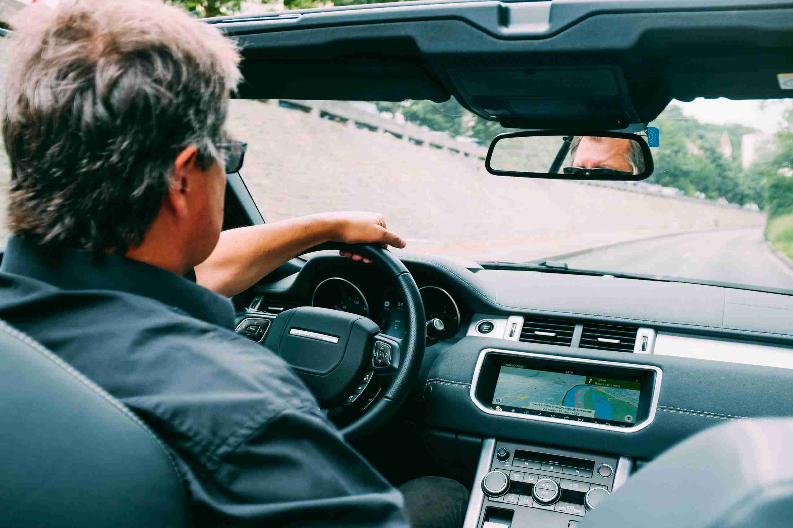 A man driving a convertible car on a scenic highway with a focus on the dashboard.