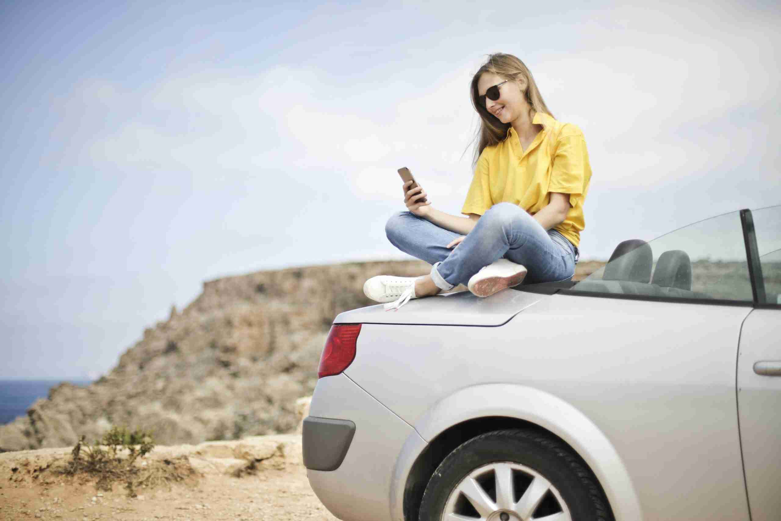 insurance crm Smiling young woman sitting on a car at the beach in Mgarr, Malta using a smartphone.